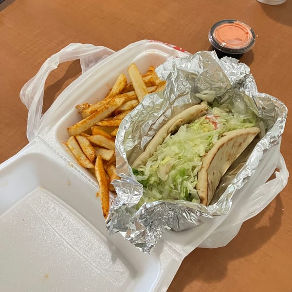 Takeout container with seasoned fries and a lettuce-filled pita wrap, accompanied by a cup of dipping sauce on a wooden table.
