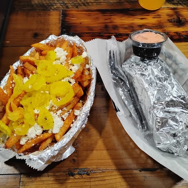 Seasoned fries topped with pickled peppers and crumbled cheese beside a foil-wrapped meal, dipping sauce, and wooden table.