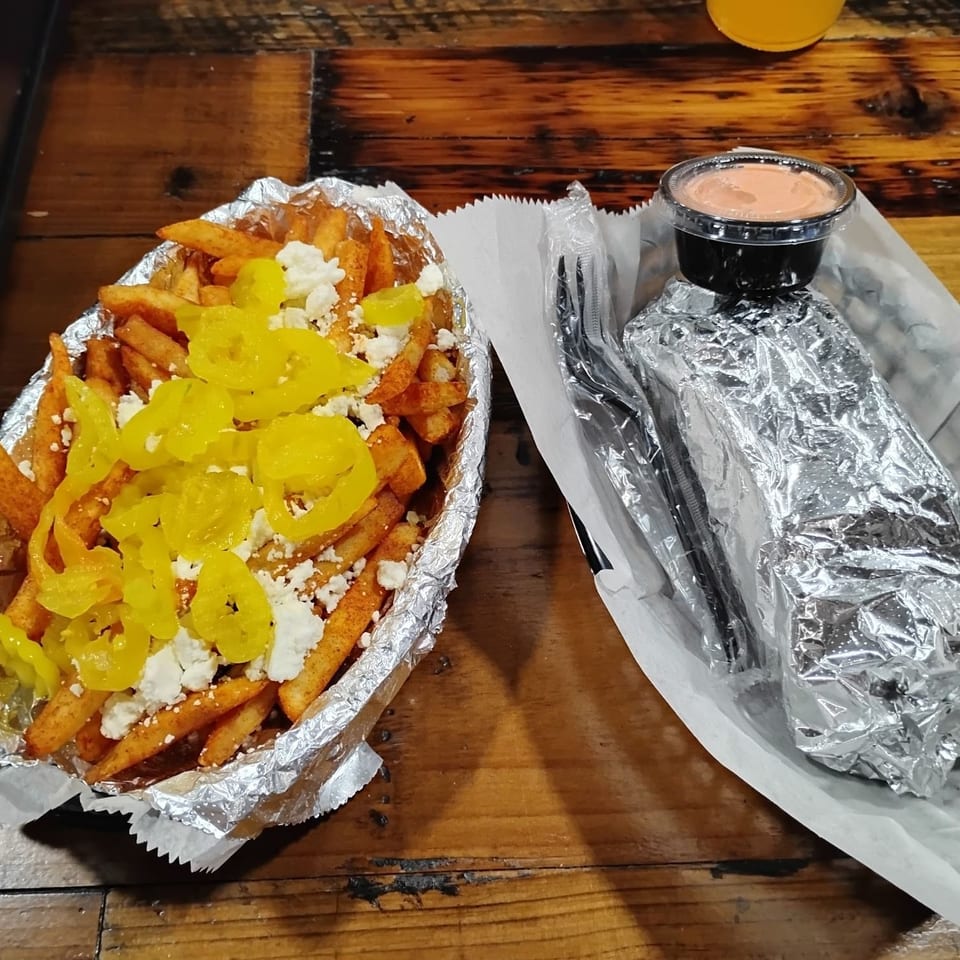 Seasoned fries topped with pickled peppers and crumbled cheese beside a foil-wrapped meal, dipping sauce, and wooden table.