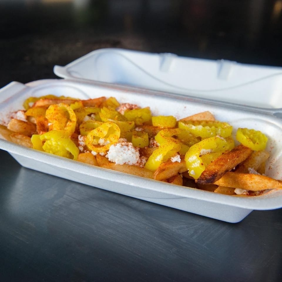 Seasoned fries topped with yellow peppers, crumbled cheese, and spices in an open foam takeout container.